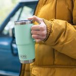 A man wearing in a yellow coat holding travel mug in his hand. A promotional drinkware travel mug customized by Faddico.