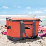 An insulated cooler bag placed on the sand by the beach, featuring a company logo for brand promotion.
