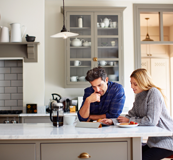 A couple is sitting in the kitchen, looking at something on their tablet.