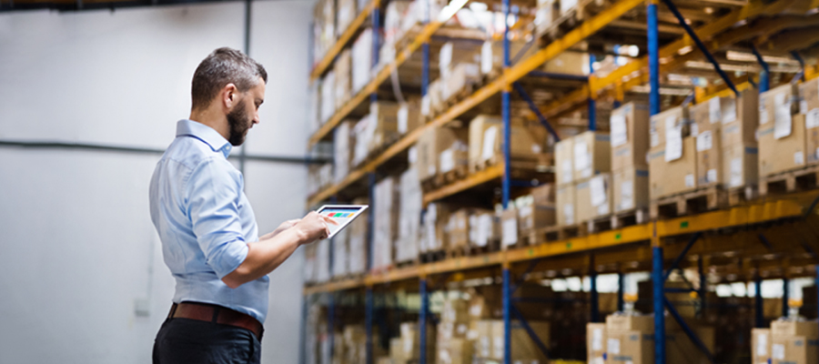 Man in a blue shirt using a tablet in a warehouse with shelves stocked with boxes - Benefits of Warehousing