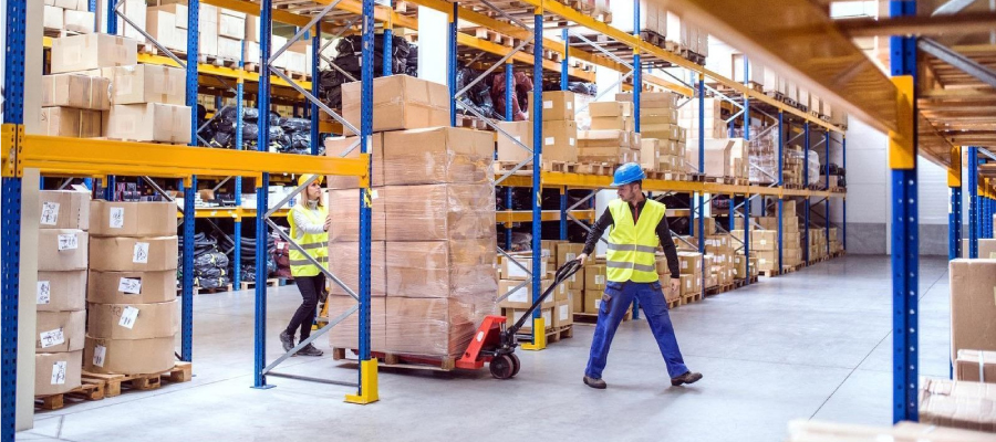 Workers in a warehouse with shelves full of boxes, one pushing a hand truck.
