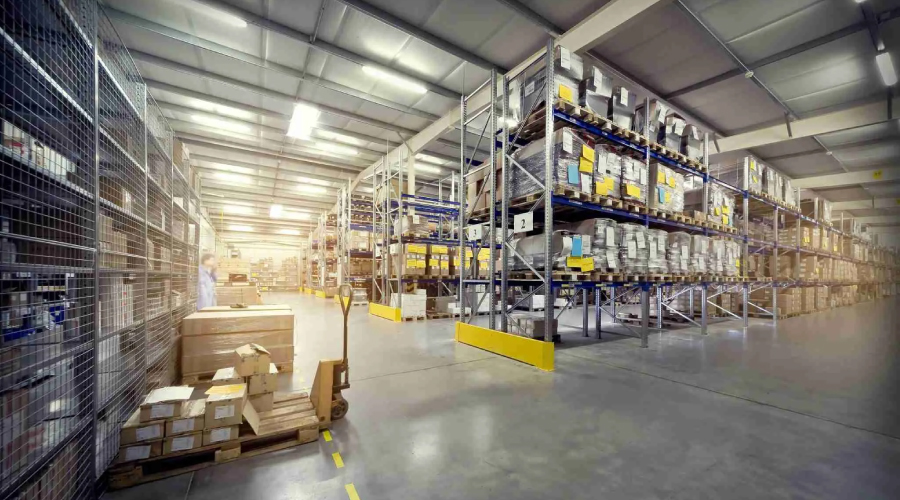 Functions of warehousing-Large industrial warehouse interior with tall shelving units stocked with various goods and a pallet jack.
